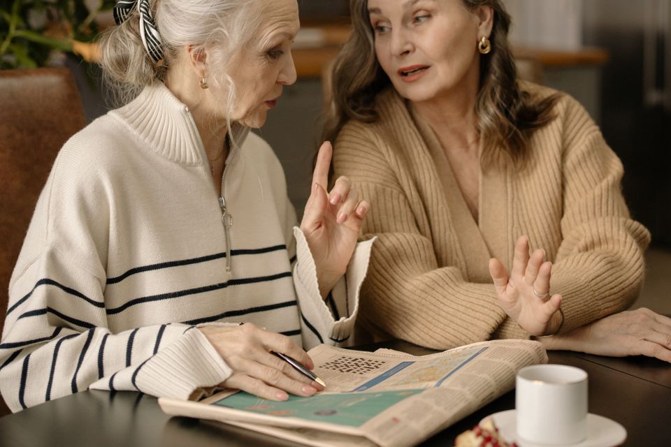 Two older women sharing conversation and a newspaper at a table